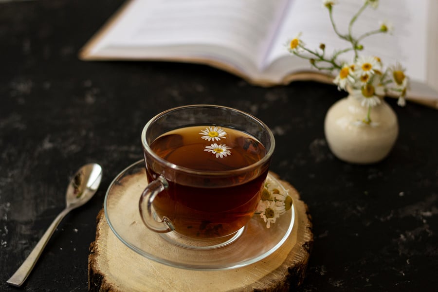 English tea with chamomile stands on a wooden frame and a teaspoon and an open book for reading lies on a black background. Nearby is a white vase with summer chamomile flowers